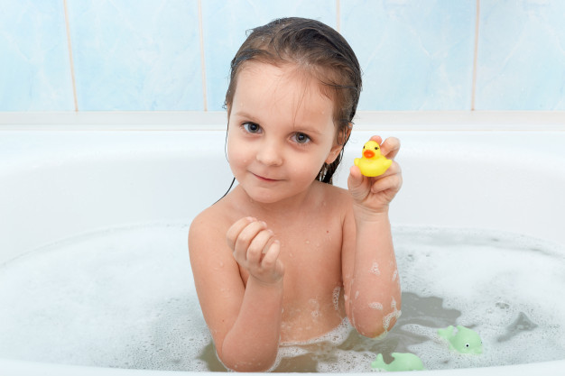 Close up portrait of happy charming little girl sitting in bath tub plays with yellow duck in bathroom. Free Photo